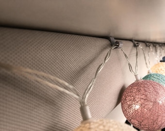 May include: A string of decorative globe lights featuring spherical shades wrapped in textured, multi-colored thread. The lights are suspended from a horizontal surface using small gray plastic clips, with visible pink, teal, and yellow spheres.