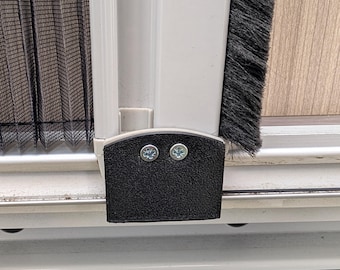 May include: Close-up of a black textured plastic component with two silver screws, attached to a white frame. A black mesh screen and a gray brush seal are visible on either side of the frame. The background shows a light wood-grain surface.