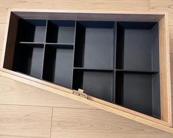 May include: A wooden drawer organizer with a light brown wood grain frame and a series of black plastic inserts. The interior is divided into seven rectangular compartments of varying sizes, arranged to fit the angled shape of the drawer frame.