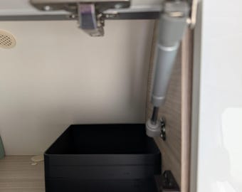 May include: A black rectangular storage bin is positioned inside a cabinet. The cabinet door is open, revealing the bin and the interior. The cabinet is made of light-colored wood. The door is held open by a silver hinge and a gas strut.