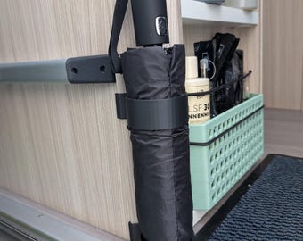 May include: A black compact umbrella secured in a holder. The umbrella is folded and has a black handle with a strap. The holder is mounted on a light-colored wooden surface. A small, black strap is attached to the umbrella handle.