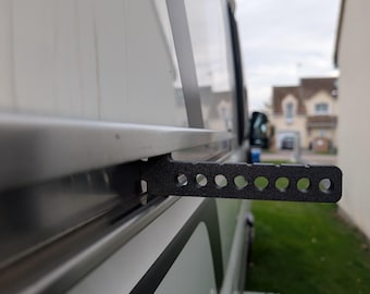 May include: A black, perforated metal bracket attached to the side of a white recreational vehicle. The bracket extends outward, with multiple circular holes along its length. The RV has a black stripe and a retractable step.