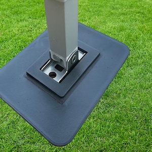 以下が含まれることがあります： テントまたはキャノピー用の濃い灰色のベースのクローズアップ。ベースは角が丸く、表面がテクスチャ加工された長方形です。銀色の金属製ブラケットがベースに取り付けられ、明るい灰色のポールを支えています。緑の芝生の上。