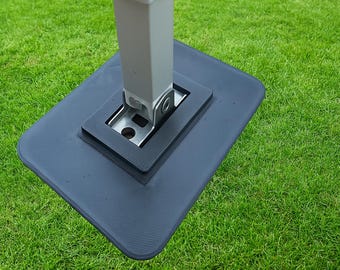 May include: A close-up of a dark gray base for a tent or canopy. The base is rectangular with rounded corners and a textured surface. A silver metal bracket is attached to the base, supporting a light gray pole. The base is on green grass.