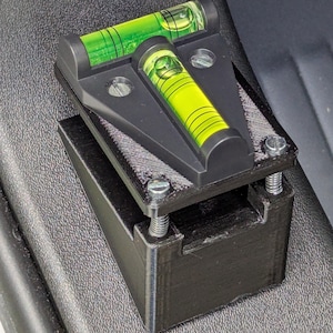 Może przedstawiać: Czarny, drukowany w 3D poziomica z dwiema zielono-żółtymi fiolkami z pęcherzykami powietrza. Poziomica jest zamontowana na czarnej podstawie i zabezpieczona srebrnymi śrubami. Fiolki wypełnione są jasnozielonym płynem.