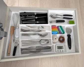 May include: A white plastic kitchen drawer organizer containing various utensils including a whisk, wooden spatula, bottle opener, knives, garlic press, peeler, forks, spoons, knives, cupcake liners, clips, twine, rubber bands, pizza cutter, and a digital timer.