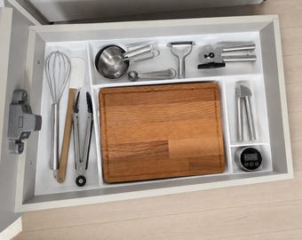 May include: A kitchen drawer organizer containing a stainless steel whisk, a silicone spatula, metal tongs, a wooden cutting board, measuring cups, a peeler, a can opener, a garlic press, and a round digital kitchen timer with a black display.