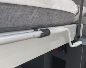 May include: Close-up of a bed frame with a gray mattress and white bedding. A white, cylindrical support pole with a black adjustment mechanism is visible. A white crank handle is attached to the frame, suggesting a lifting mechanism.