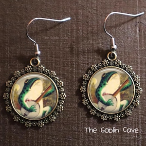 May include: A pair of silver earrings with a bronze circular pendant featuring a green frog playing a banjo. The pendant is surrounded by a decorative floral design.