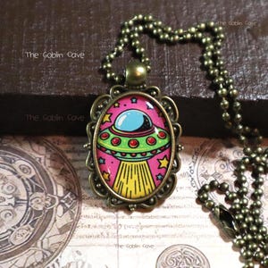 May include: Oval pendant necklace with a cartoon UFO design. The pendant has a pink background with a blue and green UFO, surrounded by stars. The chain is bronze-coloured, and the text "The Goblin Cave" is visible.