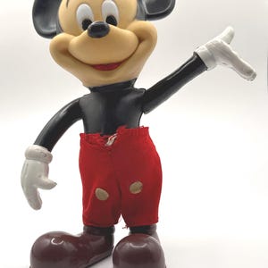 Puede incluir: Una figura vintage de Mickey Mouse. La figura tiene una cara bronceada, orejas negras y un torso negro. Viste pantalones cortos rojos, zapatos marrones y guantes blancos. La figura posa con un brazo levantado.