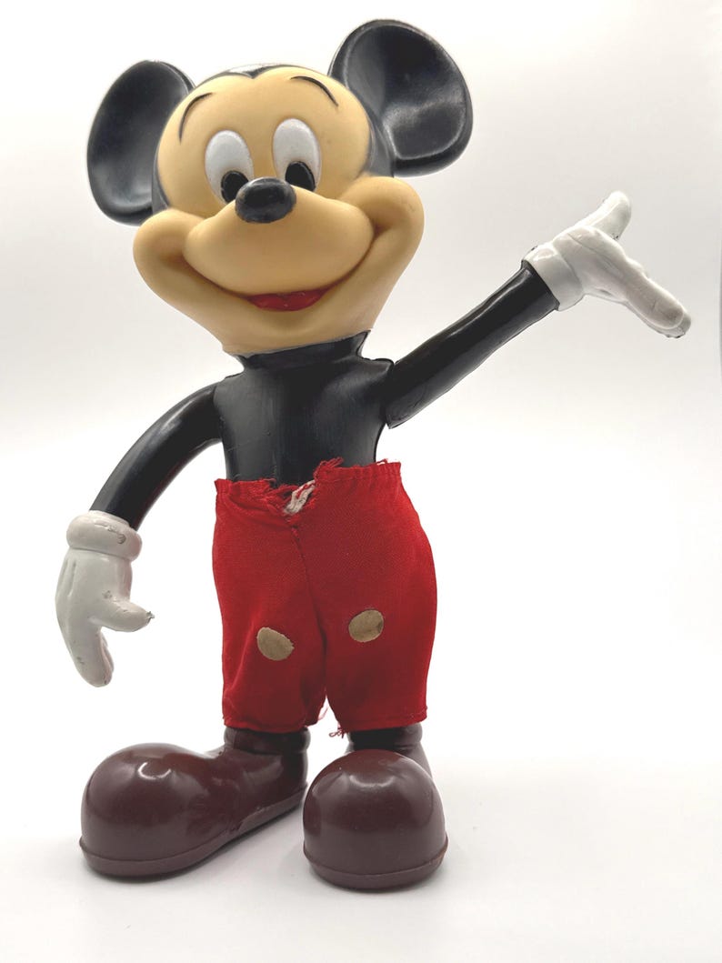 Puede incluir: Una figura vintage de Mickey Mouse. La figura tiene una cara bronceada, orejas negras y un torso negro. Viste pantalones cortos rojos, zapatos marrones y guantes blancos. La figura posa con un brazo levantado.