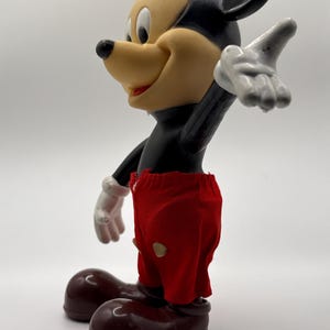 Puede incluir: Una figura vintage de Mickey Mouse. La figura tiene un cuerpo negro, una cara bronceada y grandes orejas negras. Viste pantalones cortos rojos con dos botones amarillos y zapatos marrones. La mano enguantada blanca de la figura est&aacute; levantada.