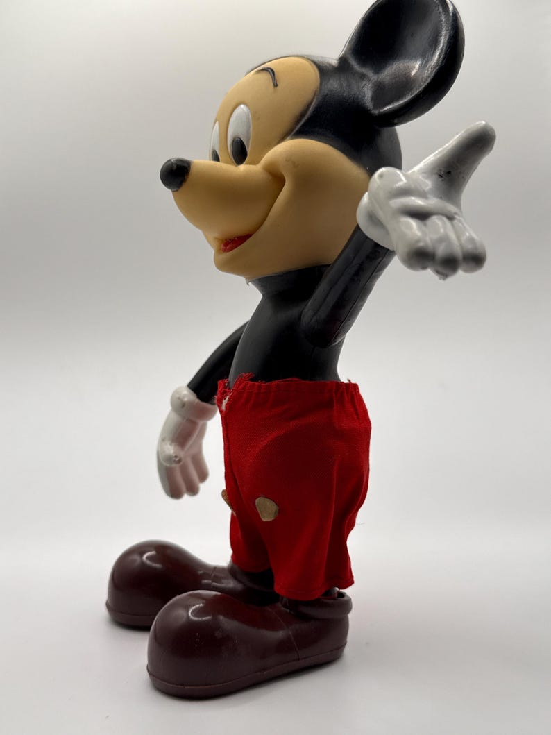 Puede incluir: Una figura vintage de Mickey Mouse. La figura tiene un cuerpo negro, una cara bronceada y grandes orejas negras. Viste pantalones cortos rojos con dos botones amarillos y zapatos marrones. La mano enguantada blanca de la figura est&aacute; levantada.