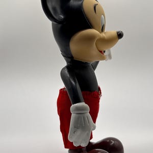 Puede incluir: Una figura vintage de Mickey Mouse. La figura tiene un cuerpo negro, grandes orejas negras y una cara bronceada con una expresi&oacute;n alegre. Mickey lleva pantalones cortos rojos, guantes blancos y zapatos marrones. La figura est&aacute; en una vista lateral.