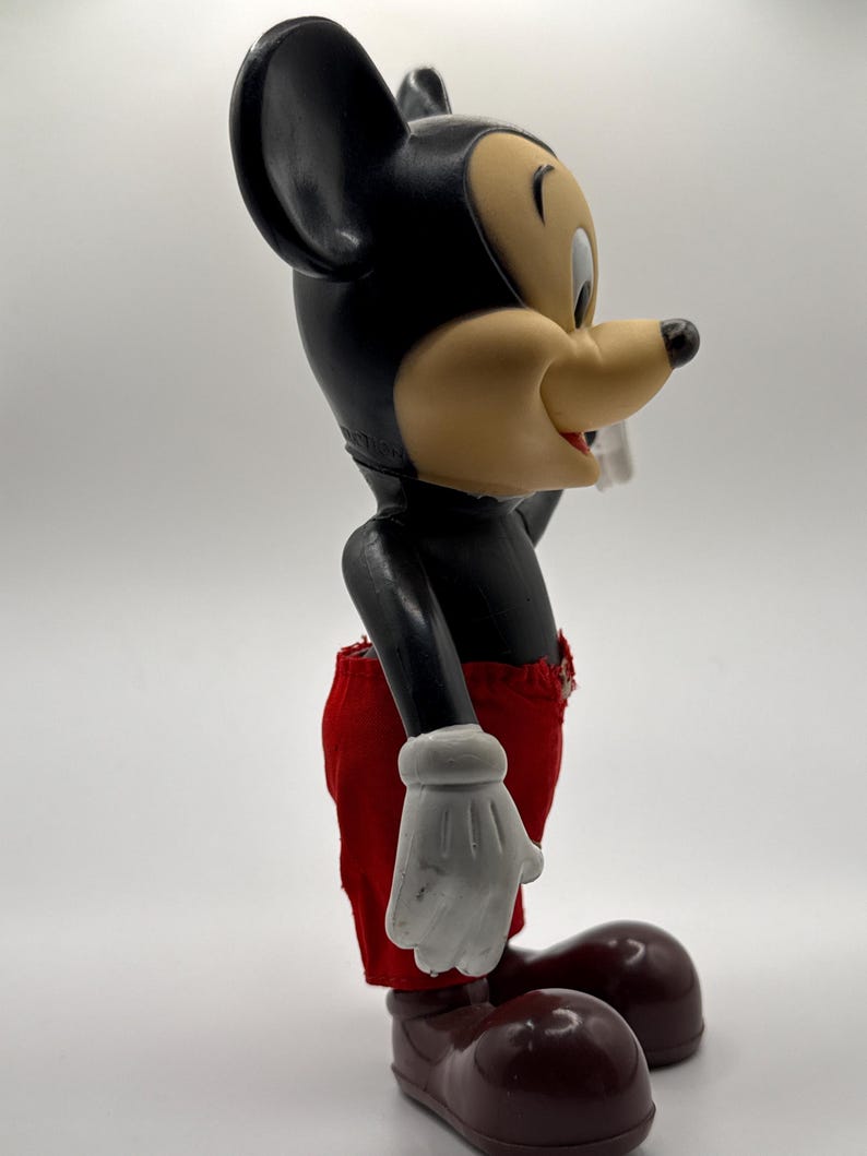 Puede incluir: Una figura vintage de Mickey Mouse. La figura tiene un cuerpo negro, grandes orejas negras y una cara bronceada con una expresi&oacute;n alegre. Mickey lleva pantalones cortos rojos, guantes blancos y zapatos marrones. La figura est&aacute; en una vista lateral.