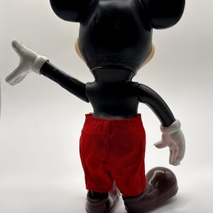Puede incluir: Figura vintage de Mickey Mouse, vista desde atr&aacute;s, con cuerpo negro, orejas grandes y pantalones cortos rojos. La figura tiene guantes blancos y zapatos marrones. El brazo est&aacute; extendido y la figura posa sobre un fondo blanco.