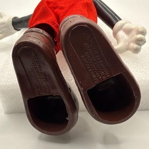 Puede incluir: Una figura vintage de Mickey Mouse, vista desde abajo, que muestra zapatos marrones y pantalones cortos rojos. El cuerpo y la cabeza negros son visibles, con guantes blancos. El texto "WALT DISNEY PRODUCTIONS R DAKIN & COMPANY PRODUCT OF HONG KONG" est&aacute; impreso en las suelas.