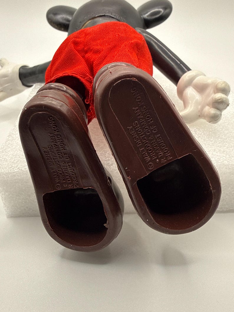 Puede incluir: Una figura vintage de Mickey Mouse, vista desde abajo, que muestra zapatos marrones y pantalones cortos rojos. El cuerpo y la cabeza negros son visibles, con guantes blancos. El texto "WALT DISNEY PRODUCTIONS R DAKIN & COMPANY PRODUCT OF HONG KONG" est&aacute; impreso en las suelas.