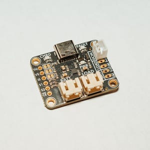 May include: A black printed circuit board featuring a USB-C port, two white battery and load connectors, and one smaller white connector. The board includes labels such as CHRG, FAULT, PWR, LOAD, BATT, and DCIN, with various gold-colored contact points.