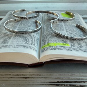 May include: An open Bible with several silver-colored, engraved bracelets. The bracelets have inspirational phrases and cross symbols. The text on the bracelets includes "FAITH, not be sight" and "Trust in the Lord with all your".