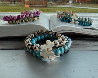 May include: Several beaded bracelets with white cross charms are displayed on an open book. The bracelets feature various colors, including turquoise, pink, and speckled patterns. The background shows a park scene with a gazebo and trees.