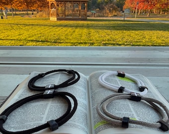 Op de afbeelding: Vier touw armbanden met een kruis bedel worden getoond op een open boek. Twee armbanden zijn zwart, een is wit en een is grijs. De achtergrond toont een park met een prieel en bomen met herfstbladeren.