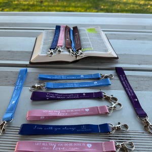 Puede incluir: Una colección de cordones de tela en varios colores, incluyendo azul, rosa y morado, con cierres metálicos plateados. Cada cordón presenta texto inspirador, como "Todas las cosas son posibles" y "Que todo lo que hagáis sea con amor."