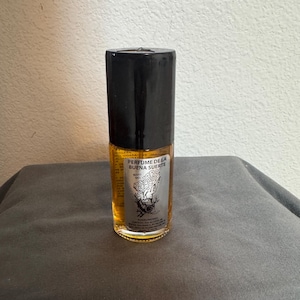 Puede incluir: Una pequeña botella de vidrio transparente con tapa negra. Contiene un líquido dorado y una etiqueta con el texto "PERFUME DE LA BUENA SUERTE" y un gráfico. La botella está sobre una superficie gris.