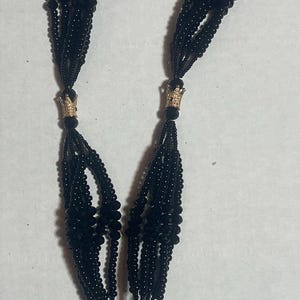 Puede incluir: Un collar de cuentas negras con múltiples hilos, con detalles de corona dorada y un colgante tallado. El collar se muestra sobre un fondo blanco, mostrando su intrincado diseño.