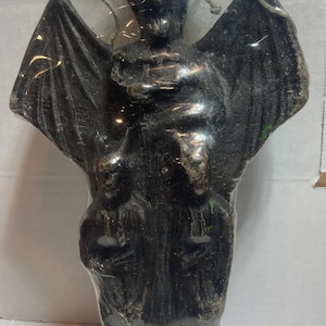 May include: A black candle shaped like a winged demon, wrapped in clear plastic. The figure has a detailed face, large wings, and is standing upright. A gold ribbon is attached to the top of the packaging.