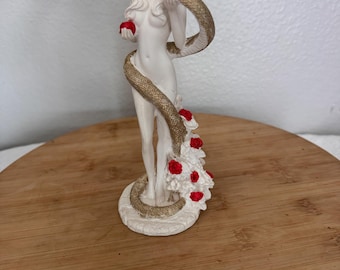 Puede incluir: Estatua blanca de una figura desnuda sosteniendo una manzana roja y una serpiente dorada. La serpiente se enrolla alrededor de la figura, y rosas rojas adornan la base. La estatua está sobre una superficie de madera.