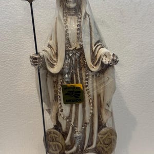 May include: A beige Santa Muerte statue with a skeletal face, wearing a hooded robe and holding a scythe. The base features the words "SANTA MUERTE" and the label "HECHO EN MEXICO".