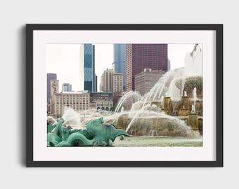 May include: A photograph of a fountain in a city with tall buildings in the background. The fountain is made of stone and has multiple tiers of water jets. The water is splashing and creating a mist. The buildings are in the background and are out of focus.