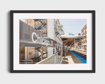 May include: A photograph of a Chicago train station platform with a sign that reads "Chicago" in white letters on a grey background. The platform is empty except for a few people walking in the distance. The platform is lined with blue tiles.