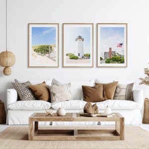 May include: Three framed photographs of a white lighthouse with a red roof, a beach scene with a wooden fence, and a red house with an American flag. The photographs are hanging on a white wall above a white couch with brown pillows.