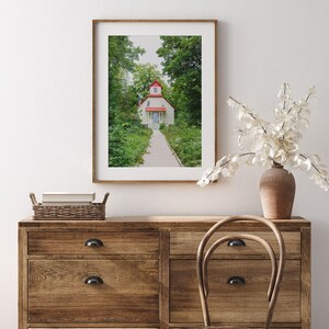 May include: A framed photograph of a white cottage with a red roof and a path leading to the front door. The cottage is surrounded by green trees and bushes.