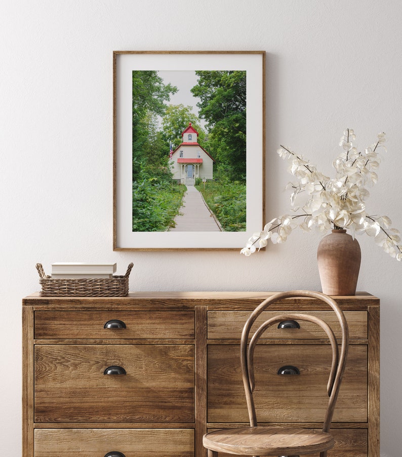 May include: A framed photograph of a white cottage with a red roof and a path leading to the front door. The cottage is surrounded by green trees and bushes.