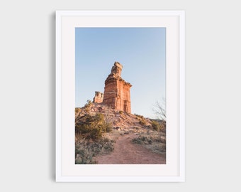 May include: A photograph of a tall, red rock formation in a desert landscape. The rock is tall and thin, with a pointed top. The rock is surrounded by a dirt path and low-lying vegetation. The sky is a clear blue.