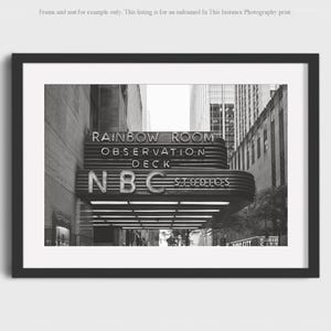 May include: Black and white photograph of the NBC Studios entrance, featuring the "Rainbow Room Observation Deck" sign. The image captures the architectural details of the building's facade, with the NBC logo prominently displayed. The photograph is framed in a black border.