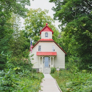 May include: A white wooden house with a red roof and a red door. The house is surrounded by green trees and bushes. A wooden walkway leads to the front door.