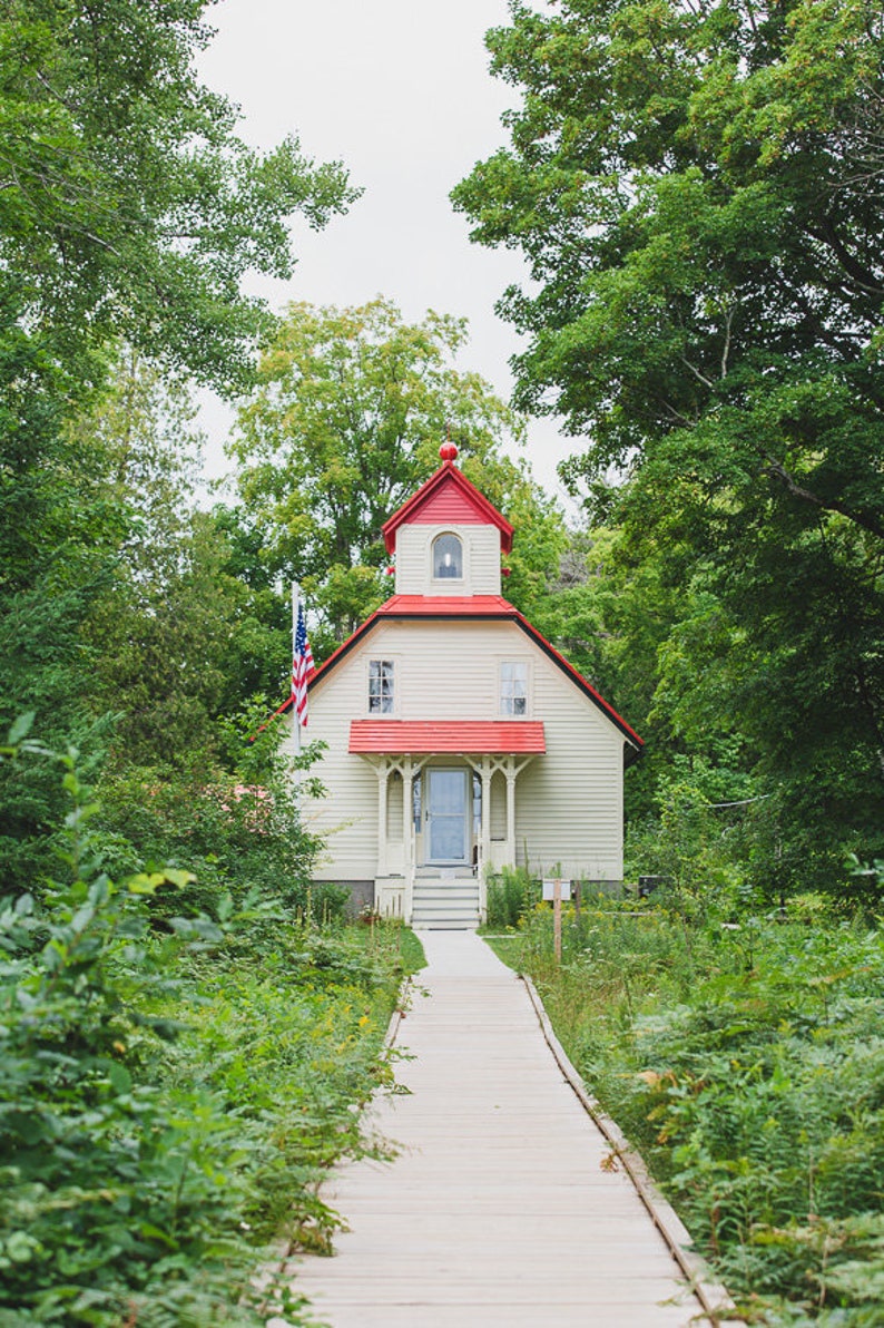 May include: A white wooden house with a red roof and a red door. The house is surrounded by green trees and bushes. A wooden walkway leads to the front door.