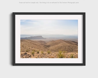 May include: A panoramic photograph of a desert landscape with rolling hills and a distant mountain range. The sky is a light blue with some clouds. The hills are covered in dry, brown grass.