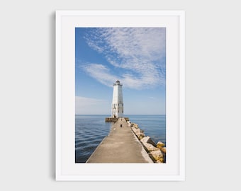 May include: A framed photograph of a white lighthouse at the end of a concrete pier extending into a calm, blue body of water. The sky is a bright blue with scattered white clouds. The photograph is in a white frame.