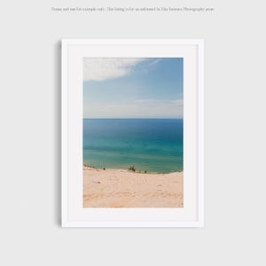 May include: A photograph of a sandy beach with a view of a blue lake. The sand is a light pink color and the water is a light blue color. The sky is a light blue color with a few white clouds.