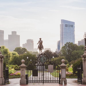 May include: A view of the Boston Public Garden, with a statue of George Washington on horseback in the foreground. The entrance to the garden is framed by two stone pillars with ornate gates. The Boston skyline is visible in the background.