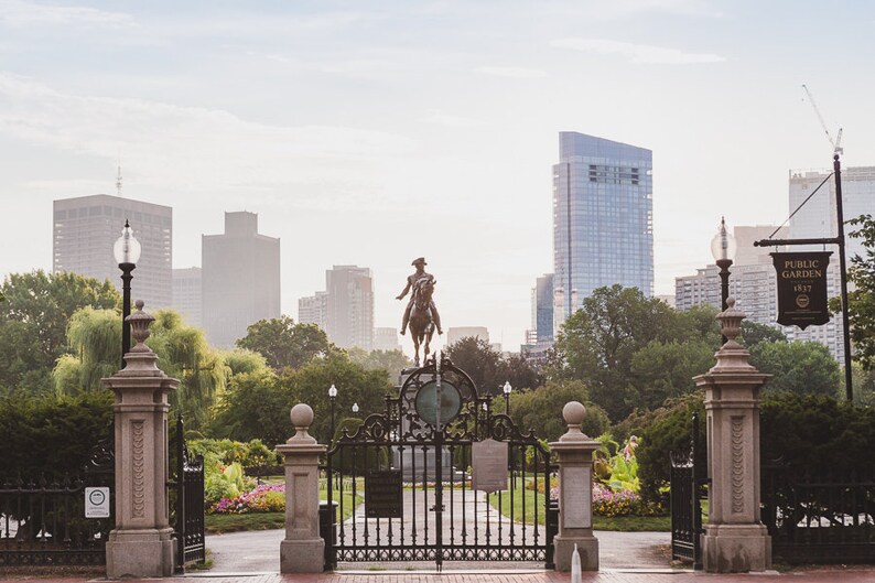 May include: A view of the Boston Public Garden, with a statue of George Washington on horseback in the foreground. The entrance to the garden is framed by two stone pillars with ornate gates. The Boston skyline is visible in the background.