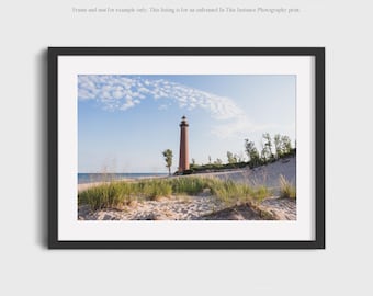 May include: A framed photograph of a tall, brick lighthouse on a sandy beach under a blue sky with scattered clouds. The lighthouse is the focal point, with dune grass in the foreground and the ocean in the background. The frame is black.