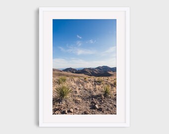May include: A photograph of a desert landscape with a blue sky and white clouds. The landscape is brown and rocky with some green bushes. The photo was taken from a hilltop looking out over the desert.