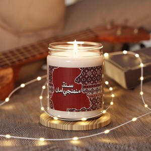 May include: A white candle in a clear glass jar with a label featuring a red map outline, intricate geometric patterns, and the Sindhi text "سند منهنجي امان". The candle sits on a circular wooden base, illuminated by a single lit wick and string lights.